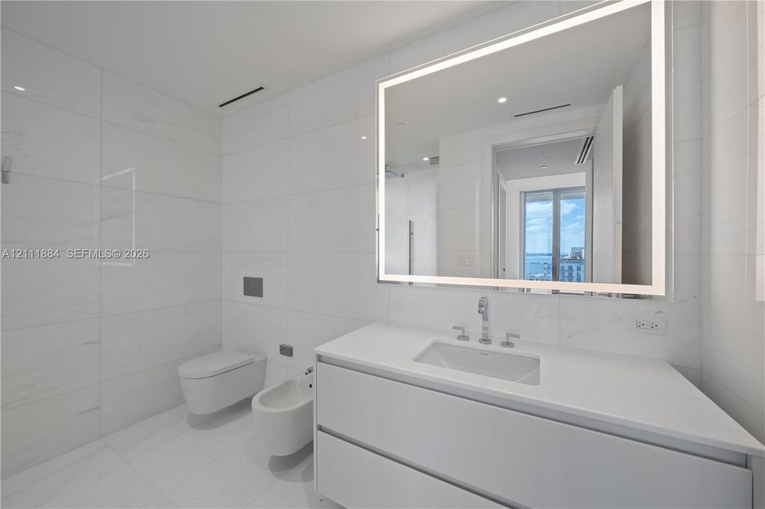 Modern bathroom featuring large-format stone-tile wall cladding, a wall-mounted toilet and bidet, a floating vanity with a stone countertop, and an integrated LED-backlit mirror