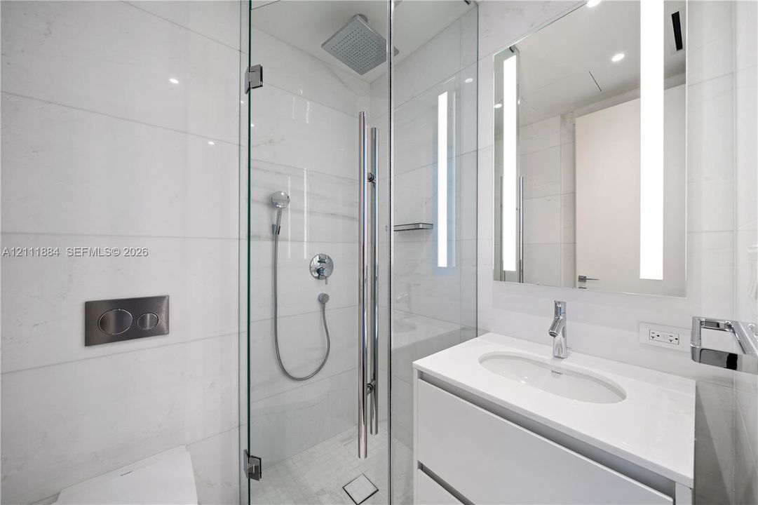 Bathroom featuring a frameless glass shower enclosure, oversized rain showerhead, wall-mounted dual-flush toilet, vanity with integrated sink, and vertical LED-integrated mirror
