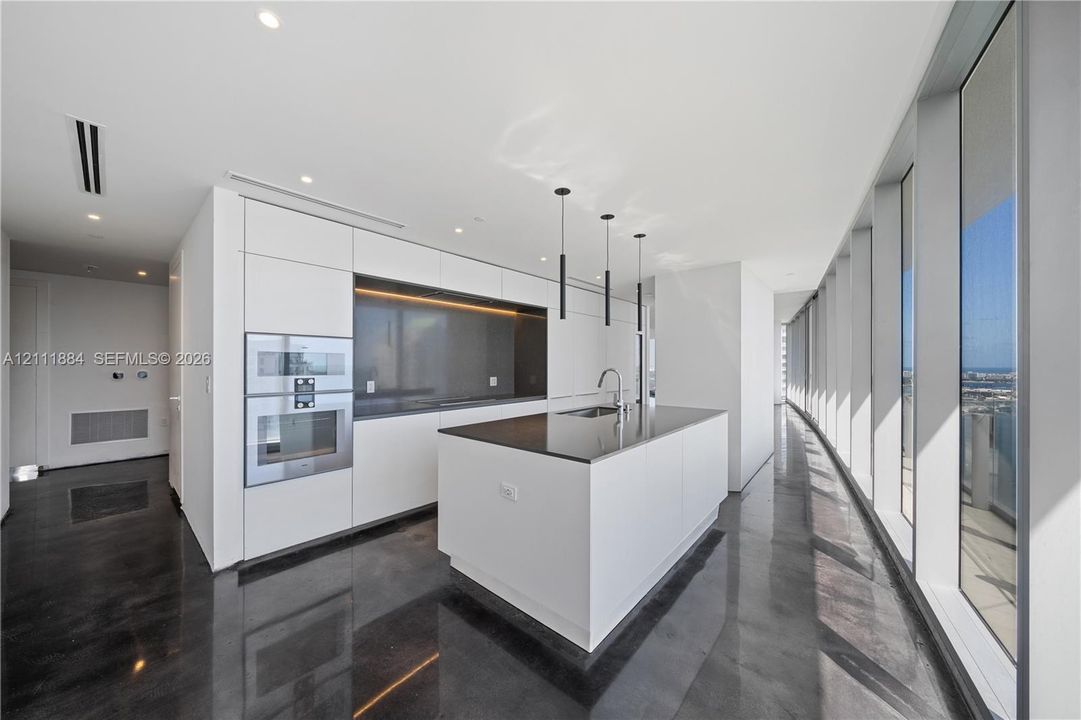 Modern kitchen featuring sleek flat-panel cabinetry, a central island with a solid-surface countertop, built-in stainless steel double ovens, floor-to-ceiling windows, and polished dark flooring