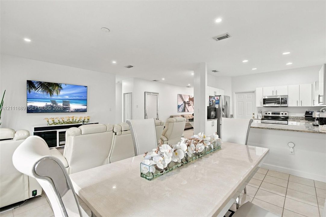 Open-concept interior featuring light-toned tile flooring, recessed ceiling lighting, a kitchen breakfast bar, white cabinetry, and stainless steel appliances
