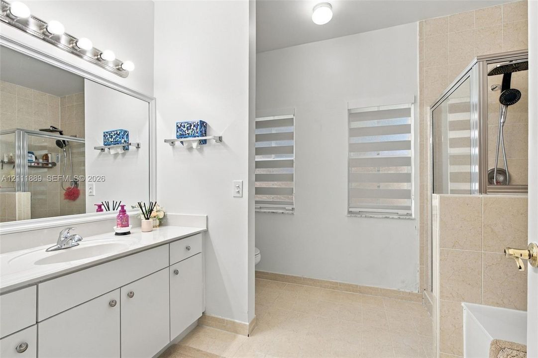 Bathroom featuring a white vanity with an integrated sink, a full-width vanity mirror, a glass-enclosed shower with tiled walls, and layered privacy blinds
