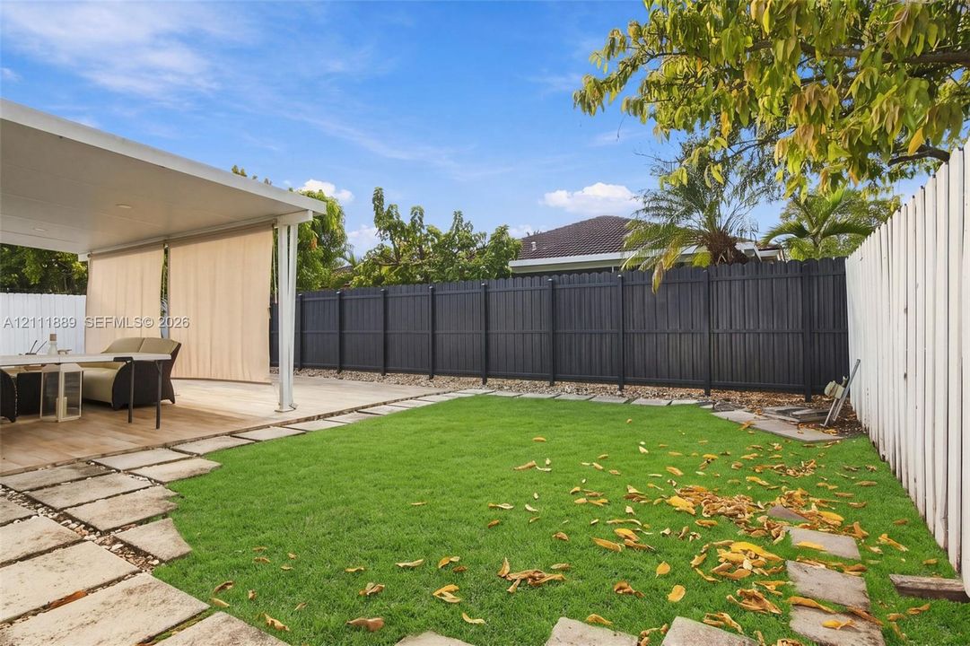 Covered patio featuring a solid roof structure and integrated privacy screens, stone paver pathway, synthetic turf lawn, and a full-height solid privacy fence