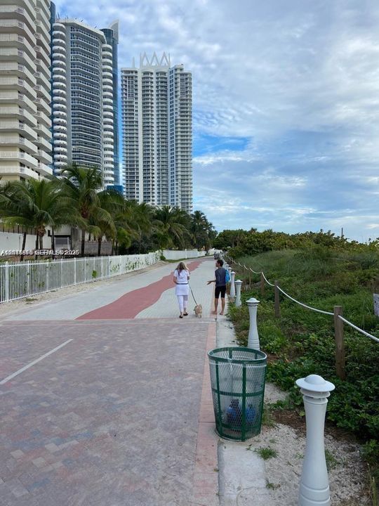 Paved pedestrian thoroughfare featuring a decorative brick-patterned walkway, white perimeter fencing, and established palm trees
