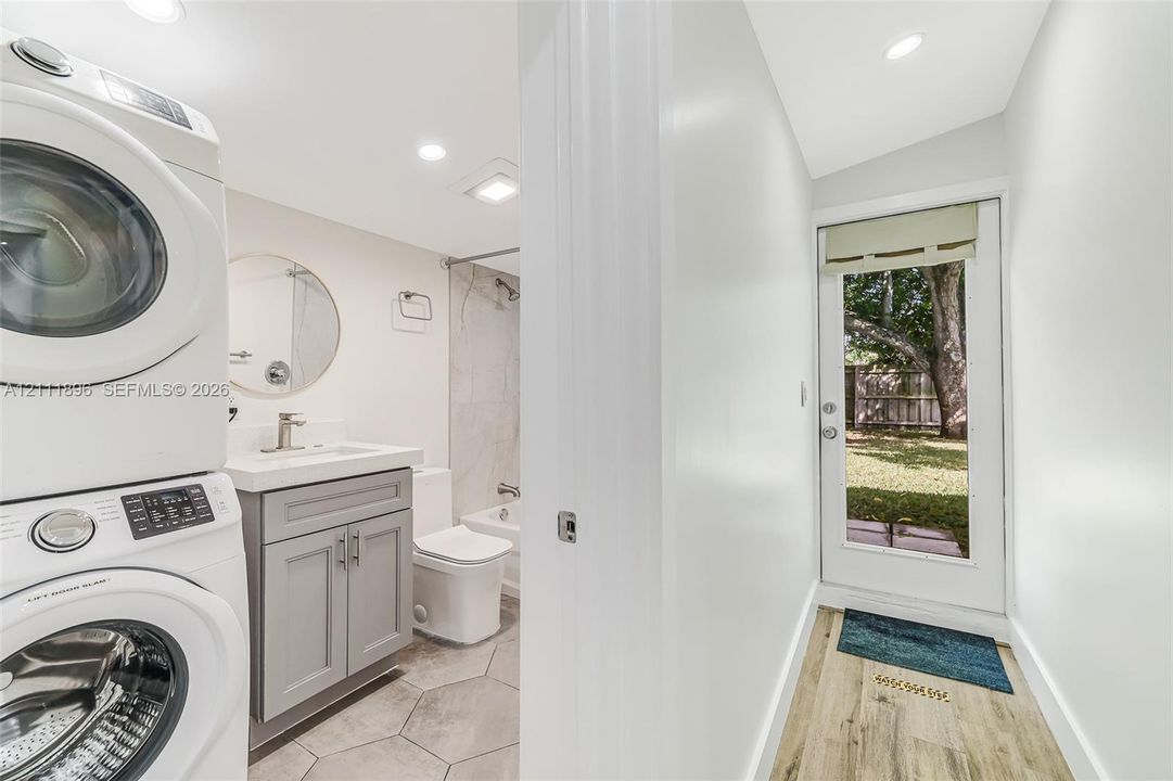 Combined utility and bathroom featuring a stacked washer and dryer, gray vanity with a rectangular basin, tile flooring, and exterior door with glass insert