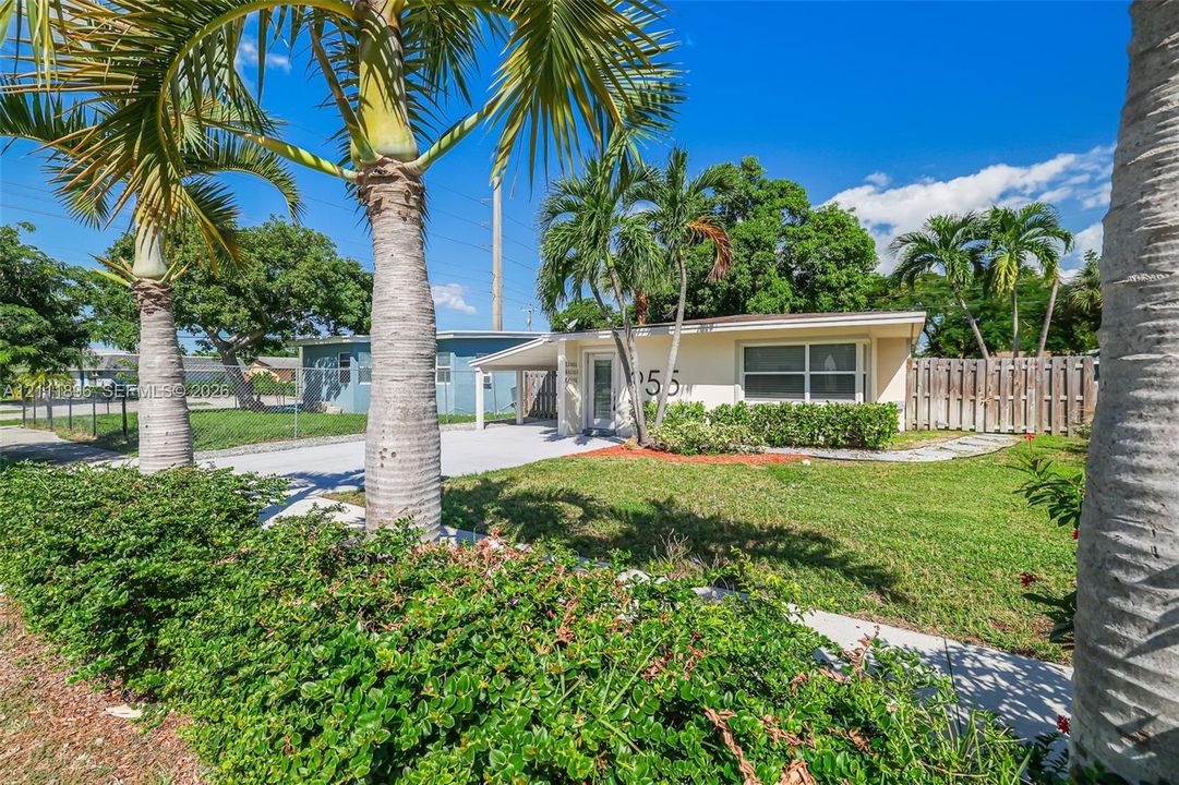 Single-story residence featuring a covered carport, expansive front lawn, mature palm trees, and wood-slat perimeter fencing
