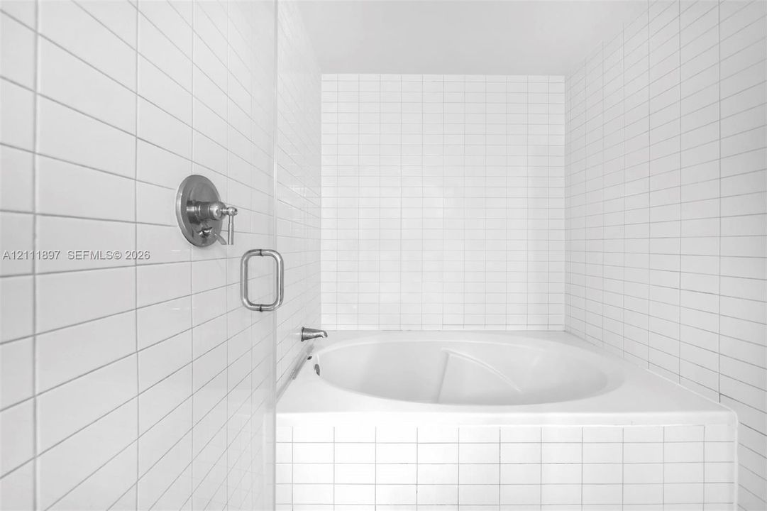 Built-in soaking tub with white square wall tile surround, complemented by rectangular subway wall tiles, a polished chrome wall-mounted faucet, and a frameless glass panel