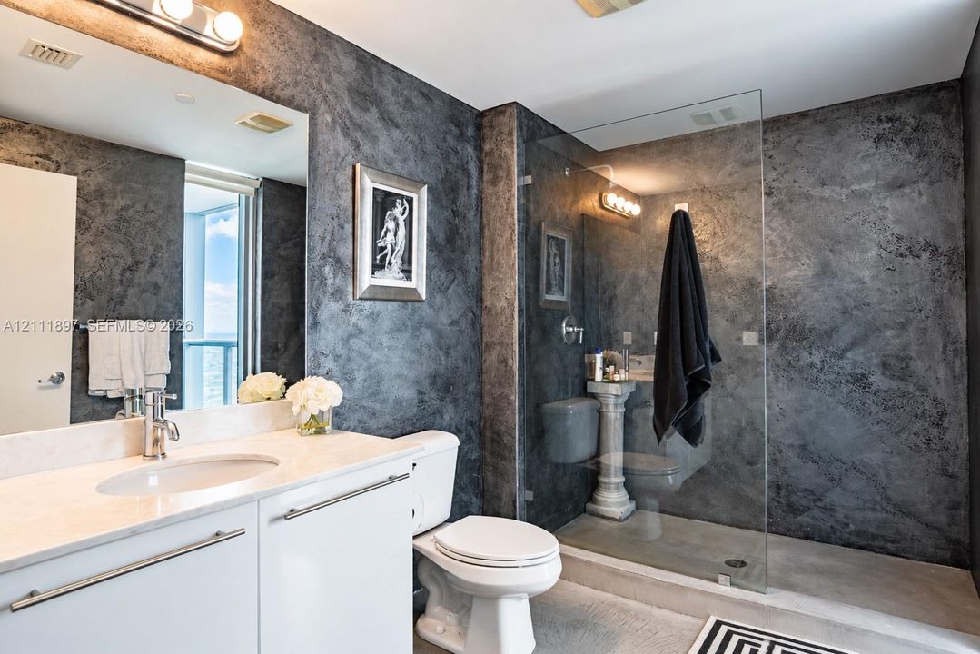 Bathroom featuring textured dark wall finish, white vanity with stone-style countertop, integrated sink, and glass-enclosed shower with pedestal fixture
