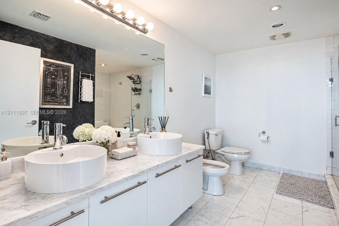 Dual vessel sinks with chrome faucets, stone-surface countertop, white cabinetry with horizontal pulls, full-width vanity mirror, and stone tile flooring