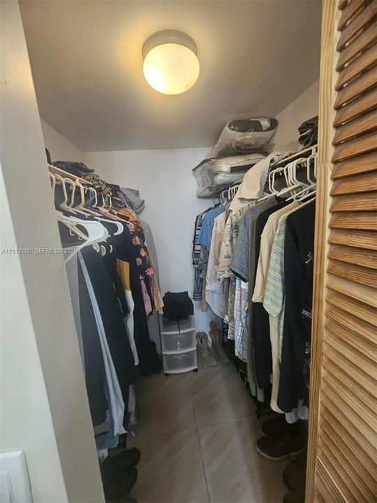 Storage closet featuring floor-to-ceiling shelving, hanging rods, tile flooring, and a flush-mount overhead light fixture, secured by wooden louvered doors
