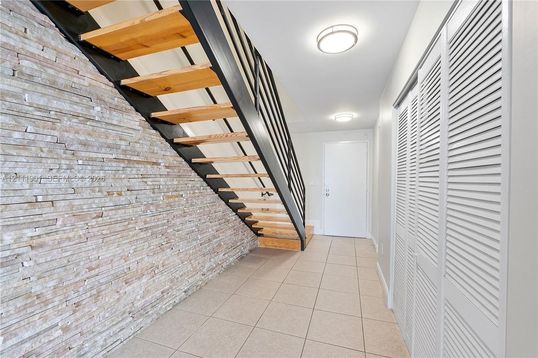 Open-riser staircase featuring natural wood treads, black metal railing, and a stacked stone accent wall, complemented by tile flooring and louvered double-door storage