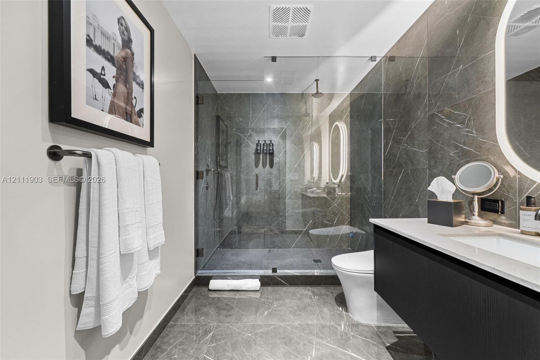 Walk-in glass-enclosed shower featuring stone-finish wall and floor tile, wall-mounted matte black towel bar, floating vanity with solid-surface countertop, and oval LED-backlit mirror