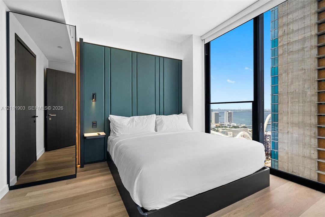 Floor-to-ceiling windows, custom teal paneling, wood-finish flooring, full-length mirror, and built-in wall-mounted bedside shelf