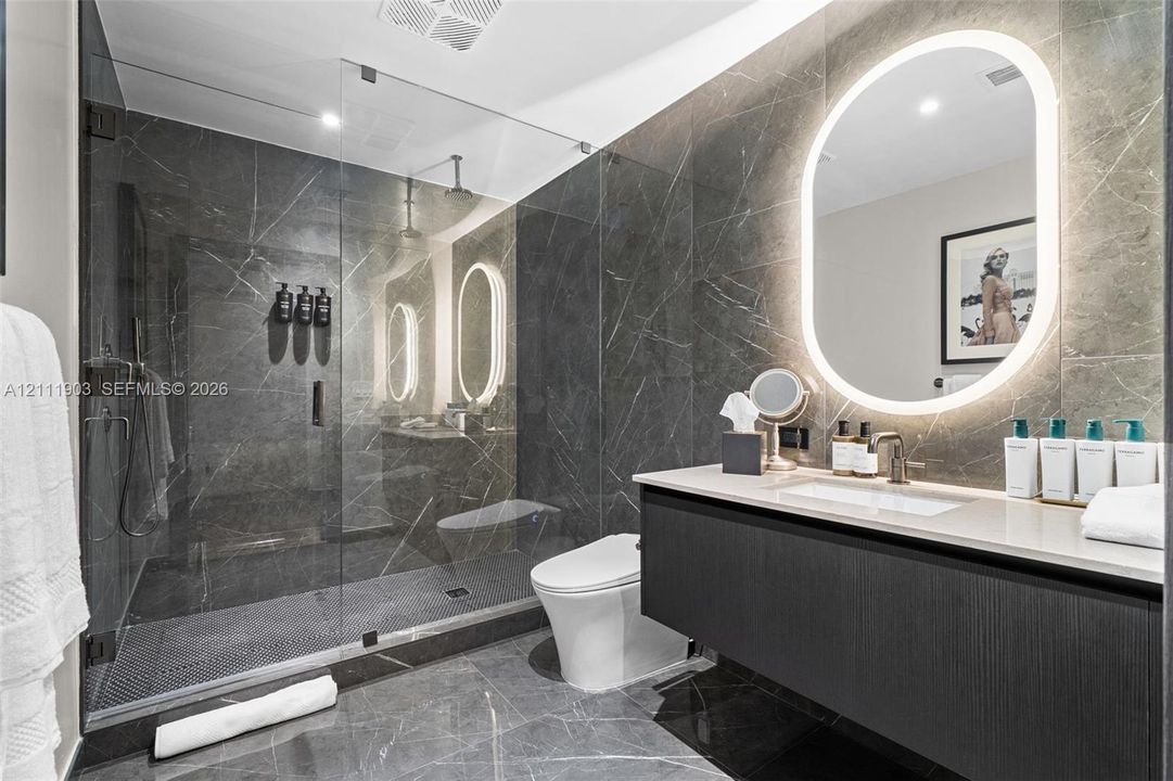 Full-height dark stone wall tiling, floating vanity with integrated countertop and sink, walk-in glass-enclosed shower with rainfall showerhead, and oval wall-mounted mirror with integrated backlighting