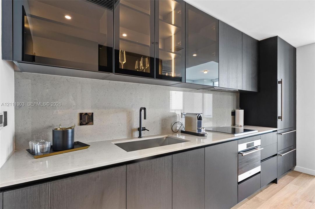 Kitchen featuring flat-panel cabinetry with glass-front upper storage, light-toned stone-surface countertops, full-height backsplash, stainless steel undermount sink with matte black faucet, and built-in appliances