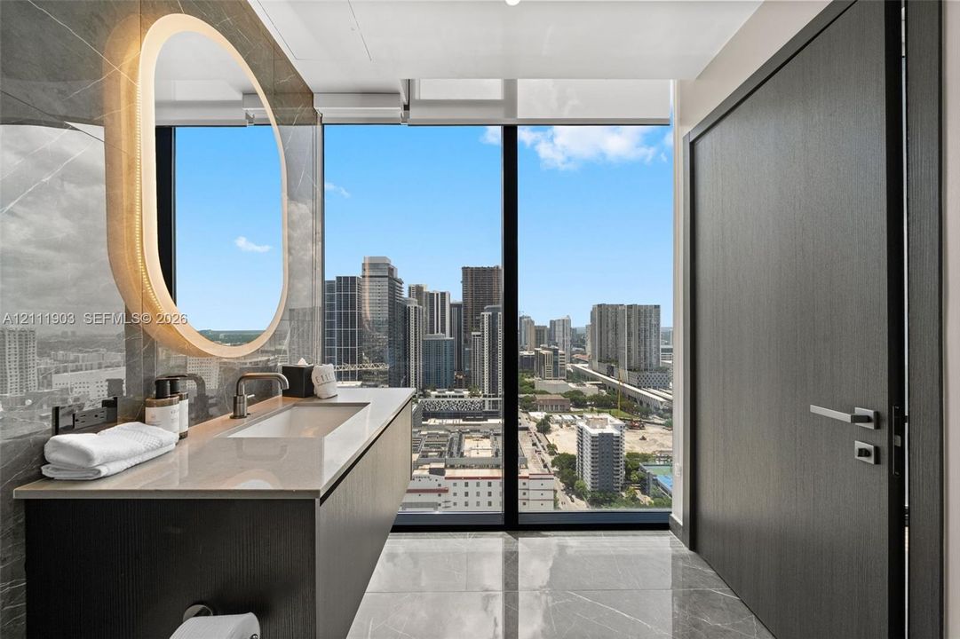 Floating vanity featuring a stone countertop, integrated sink, and wall-mounted faucet, complemented by an oval backlit mirror, full-height window, and polished stone flooring