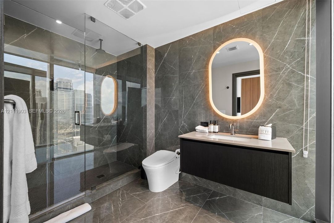 Full-height stone wall tiling, glass-enclosed walk-in shower, wall-mounted floating vanity, integrated stone countertop with undermount sink, and oval LED-backlit vanity mirror
