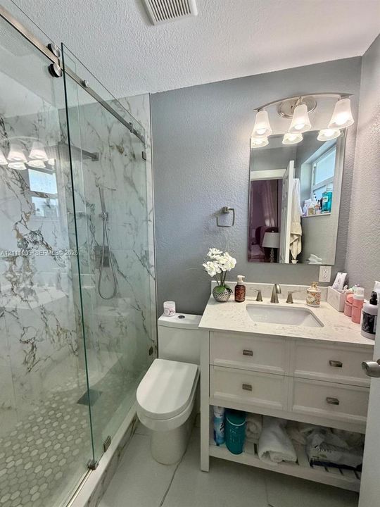 Bathroom featuring a glass-enclosed walk-in shower with floor-to-ceiling stone-finish tile, vanity with light-toned countertop, underslung sink, open lower storage shelf, and wall-mounted light fixture