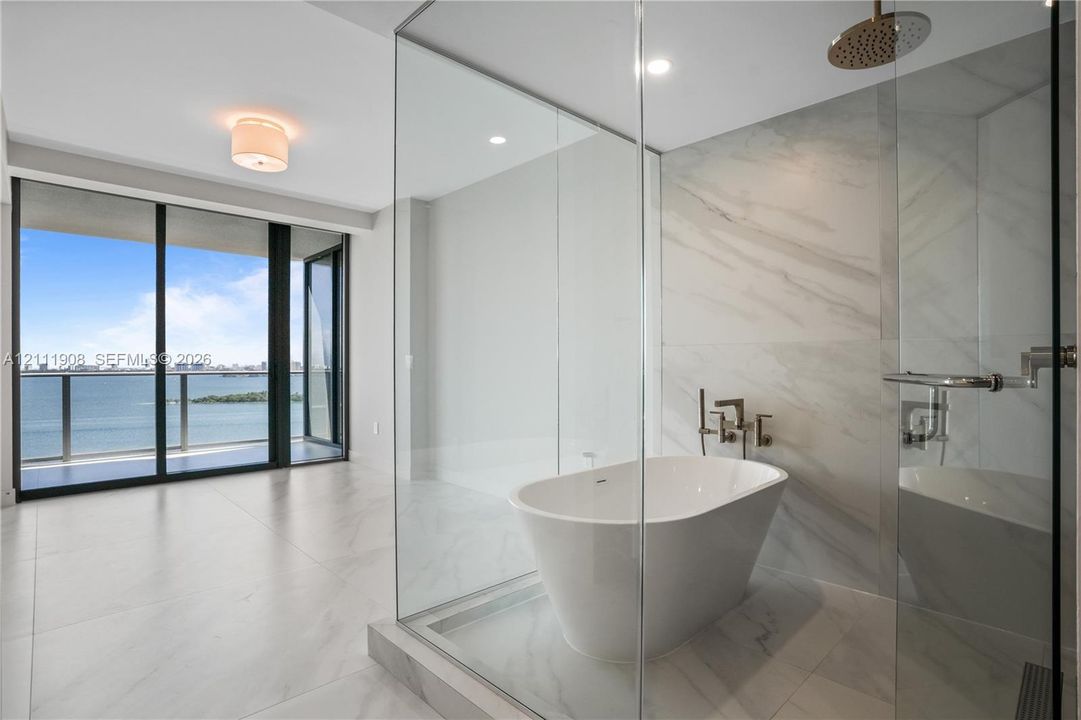 Freestanding soaking tub, glass-enclosed walk-in shower with rain showerhead, floor-to-ceiling stone-look tiling, and floor-to-ceiling windows with balcony access