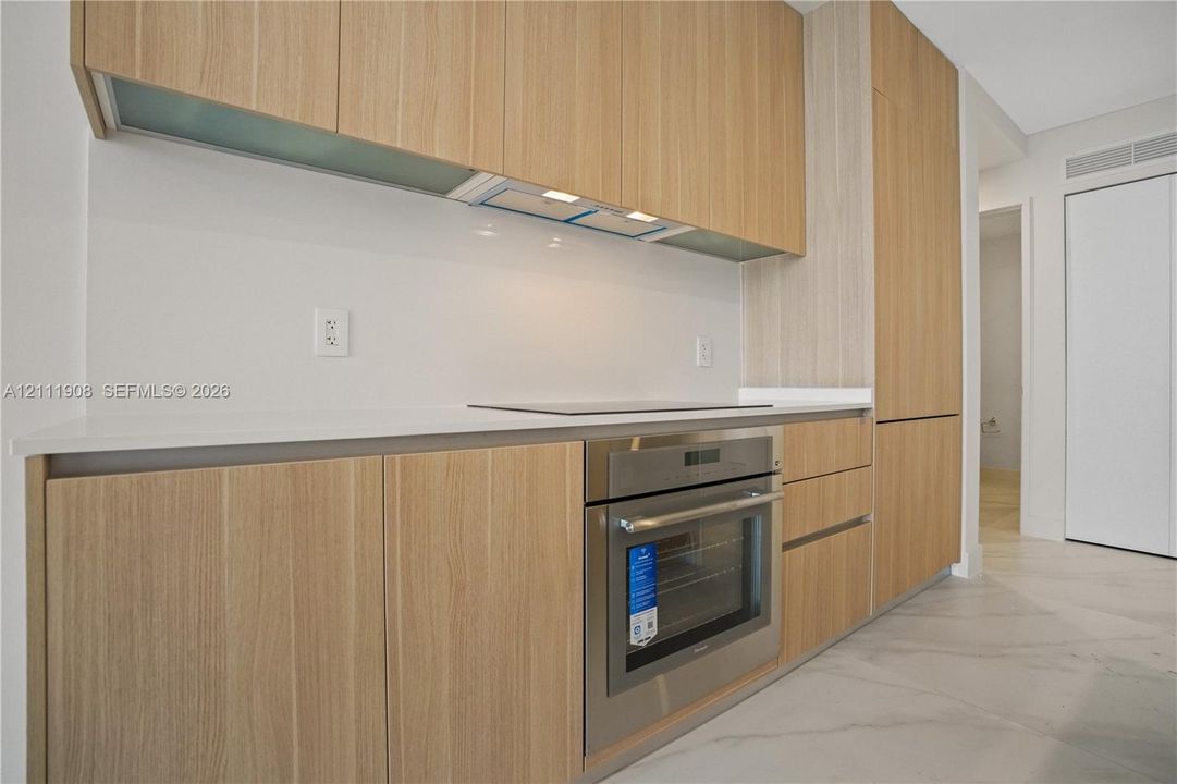 Flat-panel cabinetry with a natural wood finish, integrated stainless steel oven, cooktop, solid-surface countertops, and large-format tile flooring