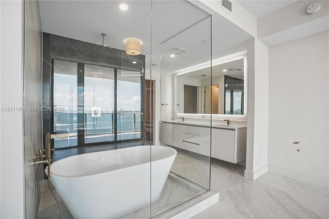 Freestanding soaking tub, glass-enclosed shower, wall-mounted double vanity, backlit mirror, and floor-to-ceiling windows