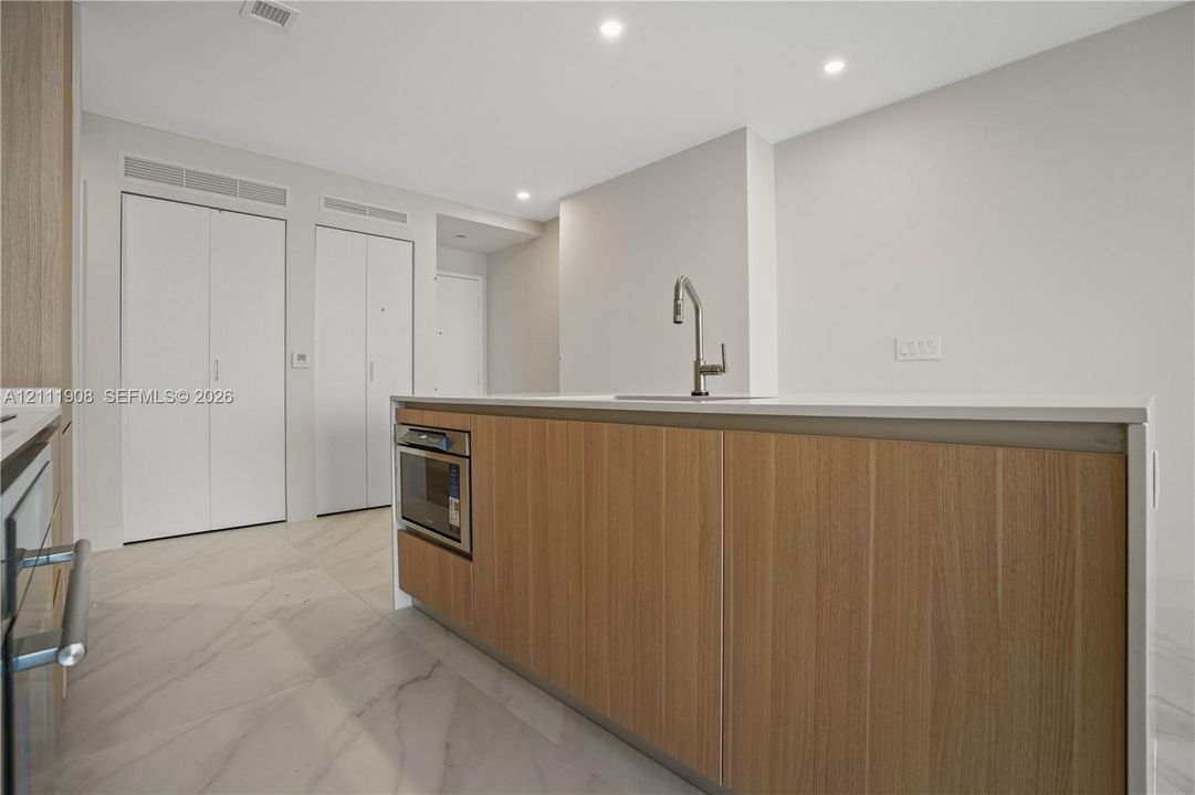 Kitchen island featuring wood-finish cabinetry, a white stone countertop, a stainless steel undermount sink, a high-arc faucet, and integrated built-in microwave, complemented by large-format tile flooring and recessed lighting