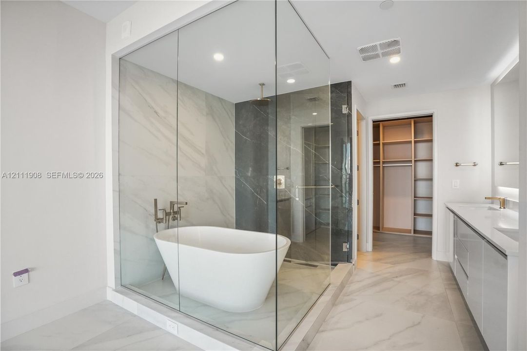 Enclosed wet room featuring a freestanding soaking tub, floor-to-ceiling glass paneling, dual-toned tiled walls, and a rainfall shower head