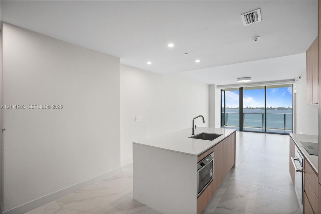 Kitchen island with stone-surface countertop, under-mount stainless steel sink, wood-finish cabinetry, floor-to-ceiling glass doors, and light-tone tile flooring