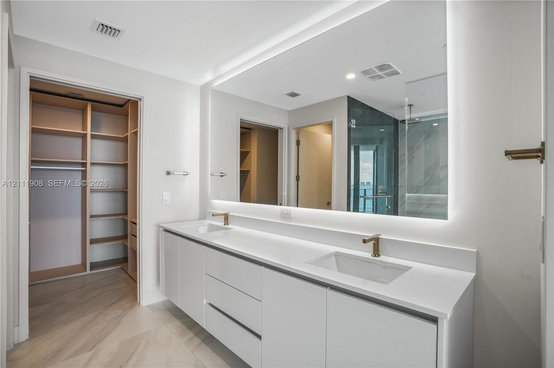 Double vanity with a composite countertop and integrated sinks, metallic hardware, full-width illuminated mirror, and custom built-in cabinetry with shelving
