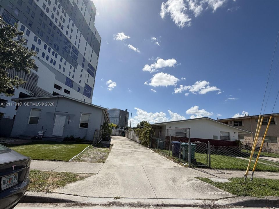 Concrete driveway providing access between two residential structures, featuring an adjacent grassy lawn area and metal chain-link perimeter fencing