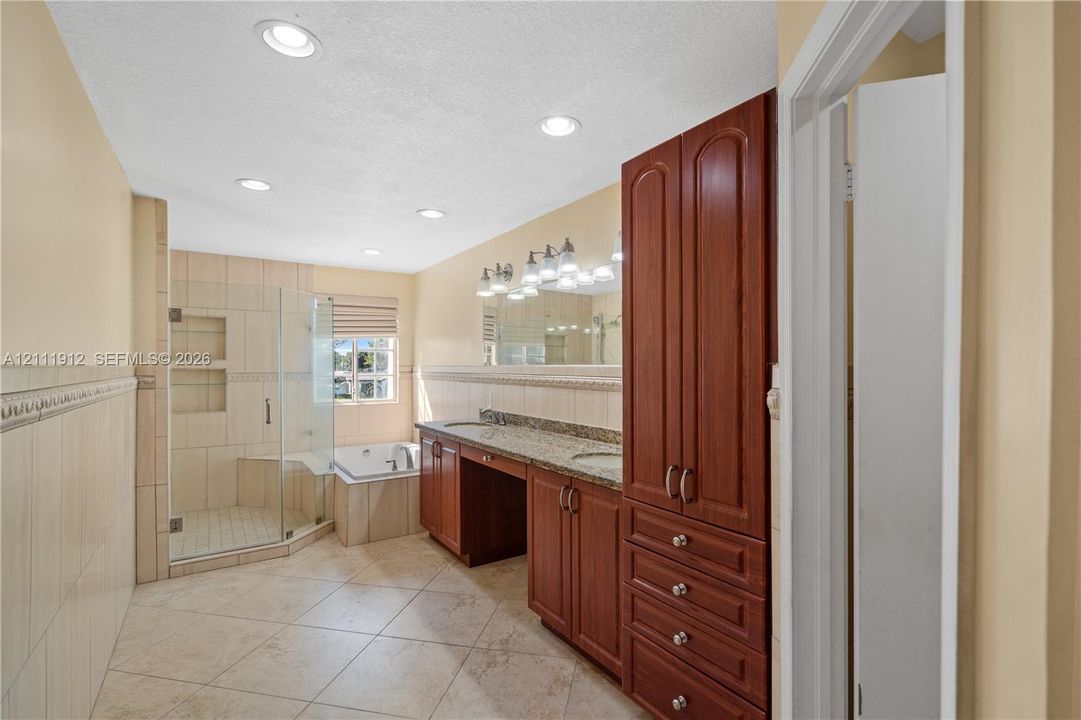 Bathroom featuring tile flooring, a dual-sink vanity with stone countertop, built-in floor-to-ceiling cabinetry, a tiled walk-in shower with glass enclosure, and a built-in soaking tub