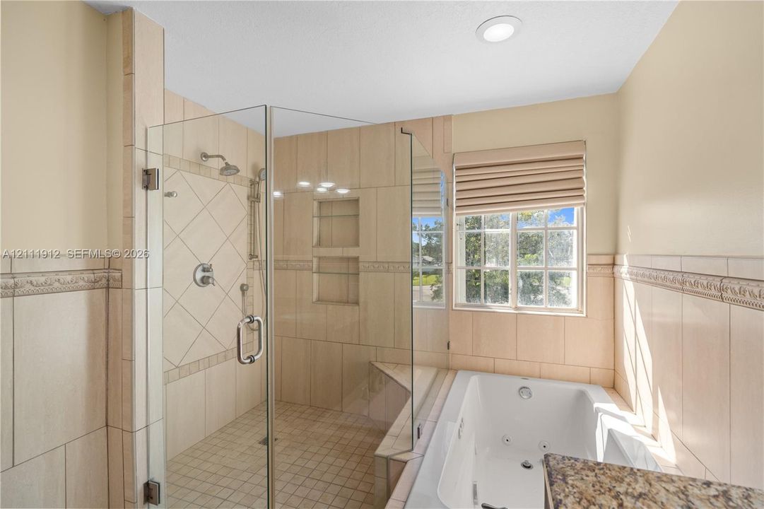 Walk-in shower with glass enclosure, decorative tile surround, built-in shelving, jetted soaking tub, and window with pleated shade