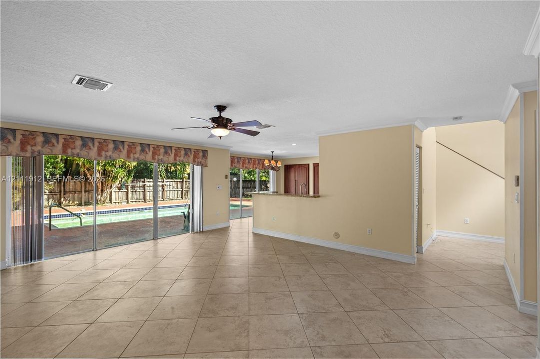 Open-concept interior featuring large-format tile flooring, crown molding, a built-in kitchen peninsula, and sliding glass doors with access to an outdoor pool area