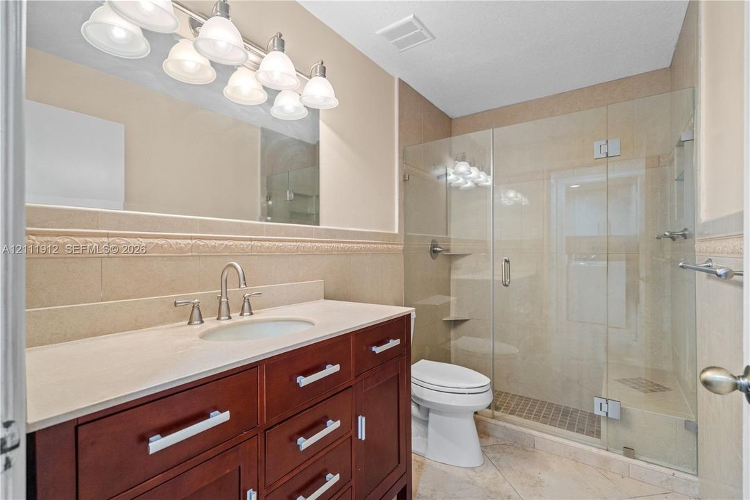 Bathroom featuring a dark wood-finish vanity, light-toned countertop with an integrated sink, frameless glass-enclosed shower, decorative tile wall border, and multi-bulb vanity lighting
