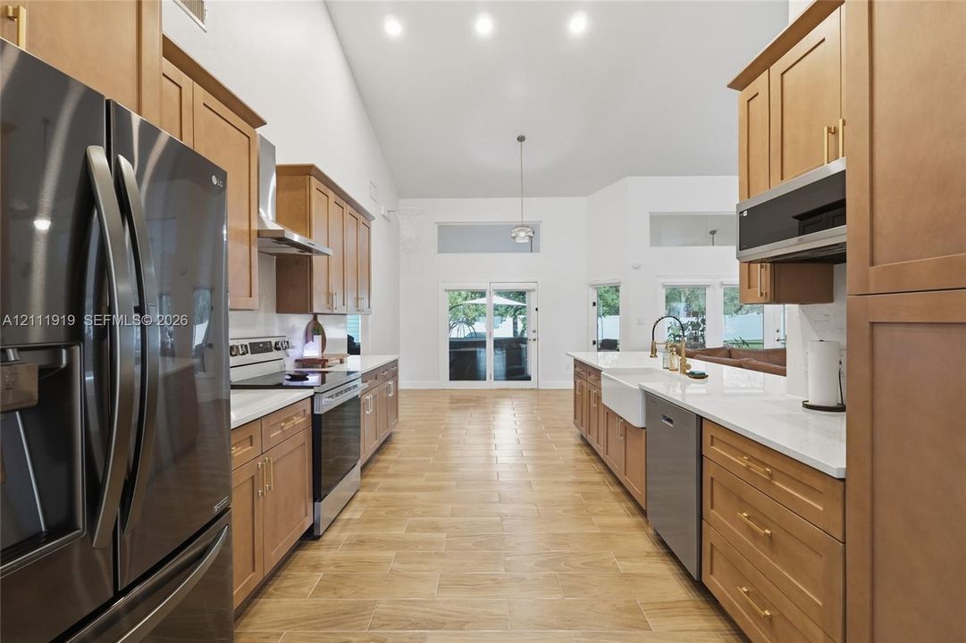 Kitchen featuring light wood cabinetry with gold-tone hardware, white countertops, a farmhouse-style sink, stainless steel appliances, and wood-finish tile flooring under a vaulted ceiling