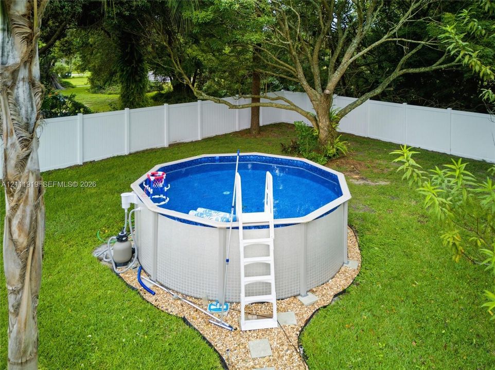 Above-ground swimming pool with surrounding gravel border, enclosed by a white vinyl privacy fence and set within a grass-covered backyard featuring mature trees
