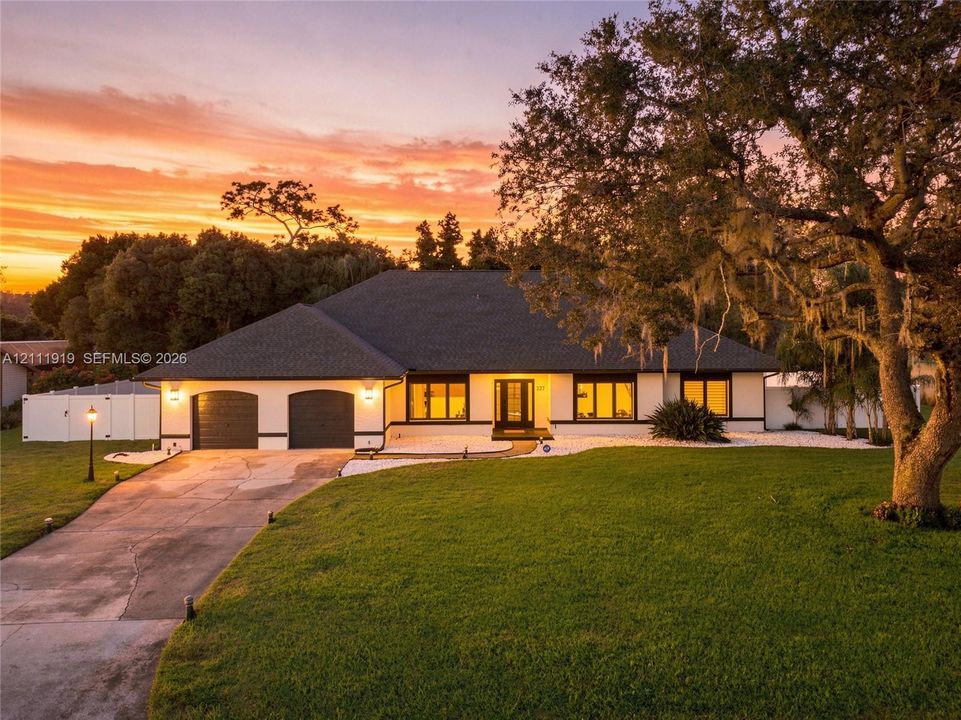 Single-story exterior featuring a hip roof, two-car garage, expansive front lawn, mature oak tree, and stone-accented landscaping