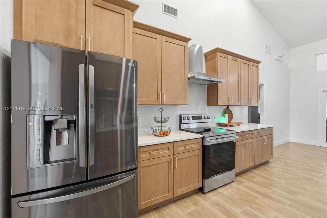 Kitchen featuring wood-finish cabinetry, light-toned countertops, stainless steel appliances, a wall-mounted chimney-style range hood, and wood-finish flooring