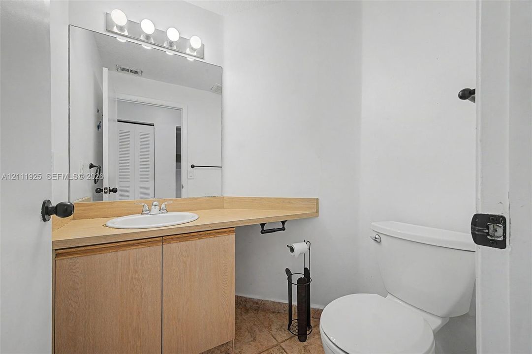 Bathroom featuring a wood-finish vanity cabinet, light-toned countertop with an integrated basin, wall-mounted mirror, four-bulb vanity light fixture, and tile flooring