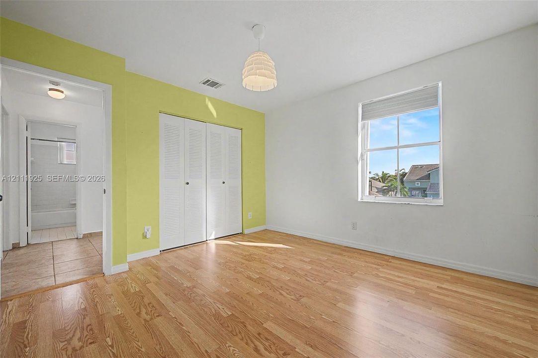 Wood-finish flooring, double-door louvered closet, pendant lighting, and single-hung window with roller shade
