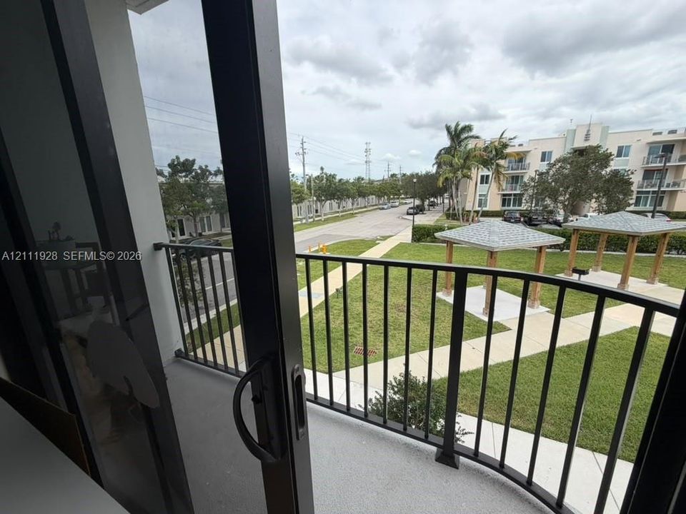 Balcony with black metal railing and concrete flooring, overlooking a green space featuring paved walkways and wooden-frame gazebos