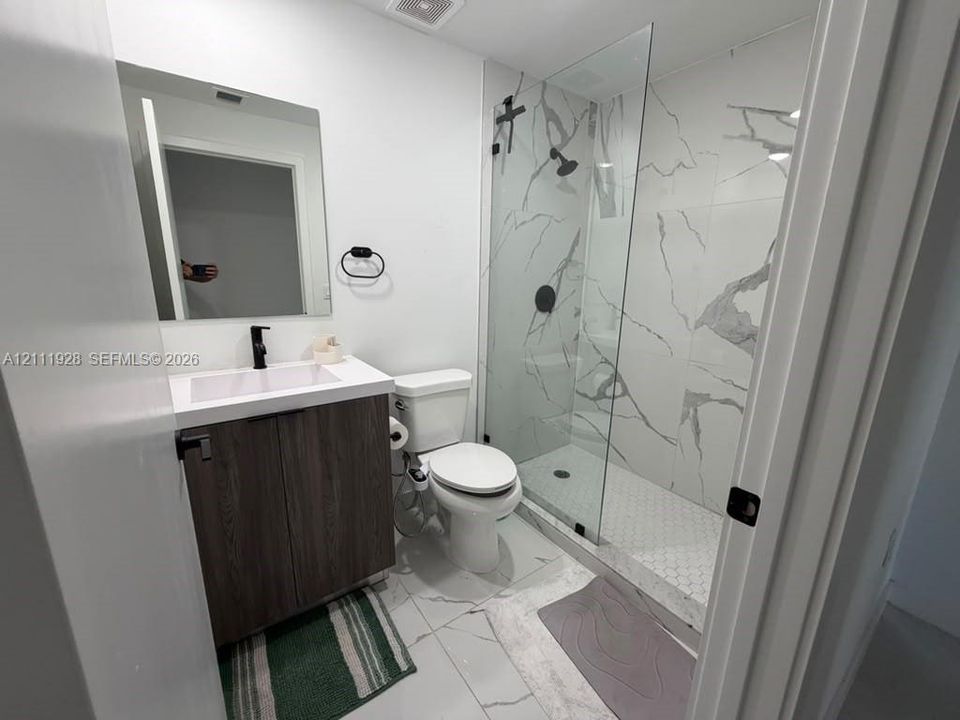 Modern bathroom featuring a wood-finish vanity, white countertop with integrated sink, walk-in shower with floor-to-ceiling veined tile, glass enclosure, and matte black fixtures