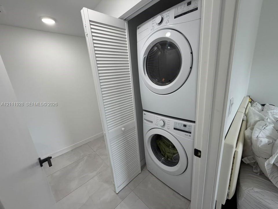 Stacked white front-load washer and dryer unit enclosed by white louvered bifold doors, featuring tile flooring and recessed ceiling lighting
