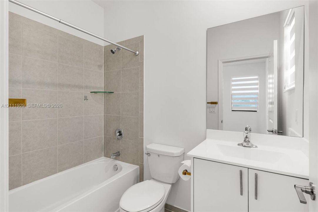 Bathroom featuring a white vanity with a composite countertop, a large rectangular wall-mounted mirror, a tub-shower combination with neutral tile surround, and gold-finish hardware