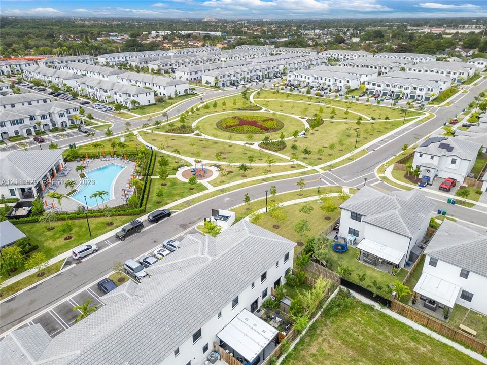 Aerial perspective of a residential community featuring a central circular garden, a swimming pool, a playground, and paved streets