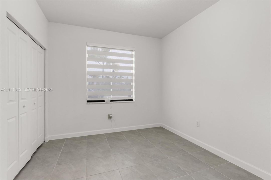 Neutral-toned ceramic tile flooring, built-in closet with multi-panel folding doors, and a window featuring adjustable horizontal light-filtering shades