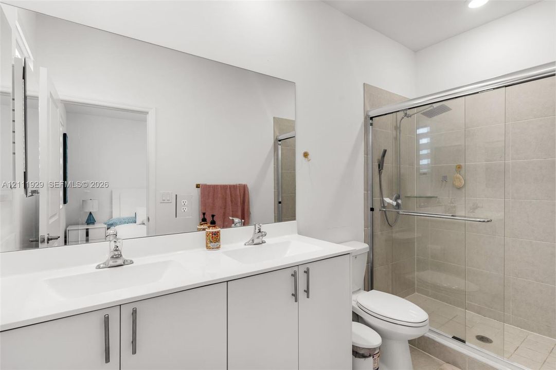 Double vanity featuring a solid-surface countertop with integrated sinks, frameless full-width vanity mirror, white cabinetry with brushed-metal hardware, and a glass-enclosed shower with tiled surround