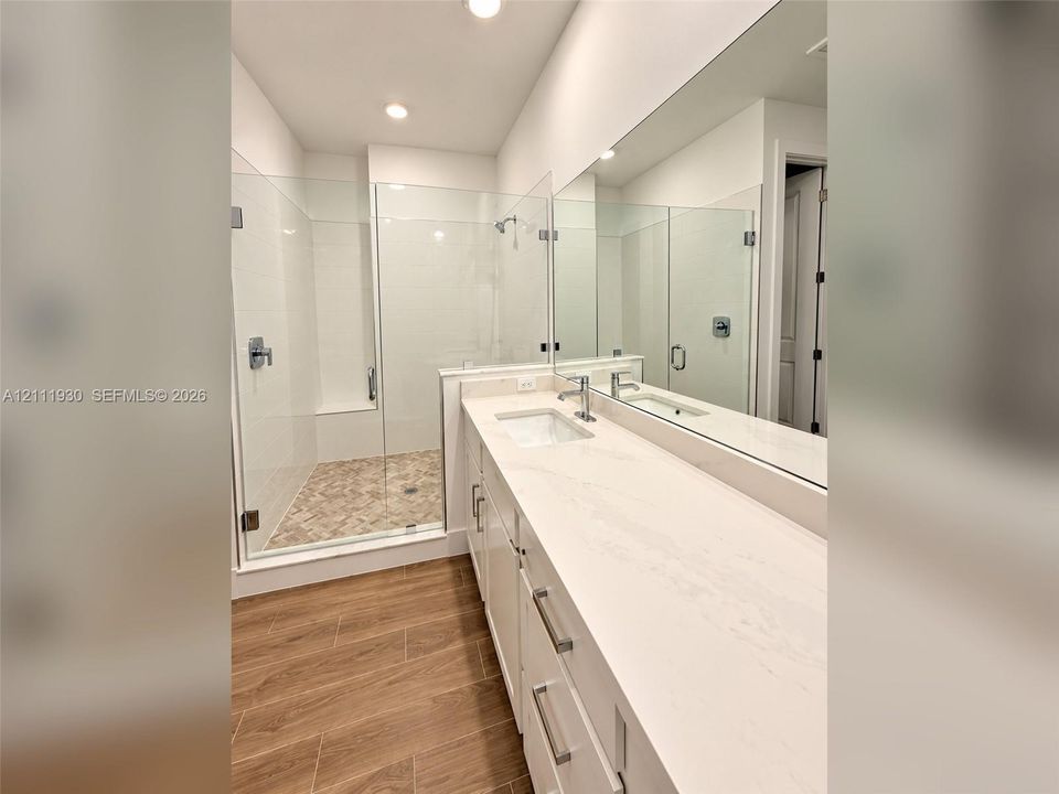 Bathroom featuring a walk-in shower with glass enclosure, light-toned stone-finish shower flooring, white cabinetry with countertop, full-width vanity mirror, and wood-finish flooring