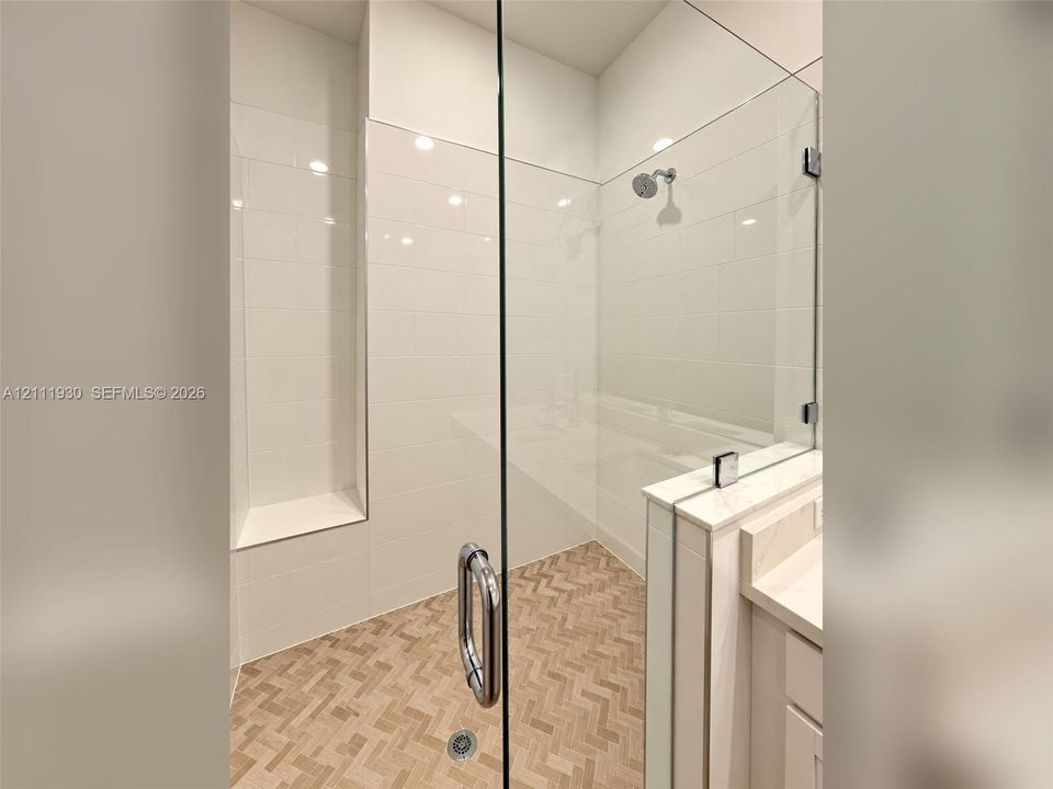 Walk-in shower featuring a frameless glass door, herringbone-pattern tile flooring, white wall tiling, built-in recessed shelving, and a metal shower head