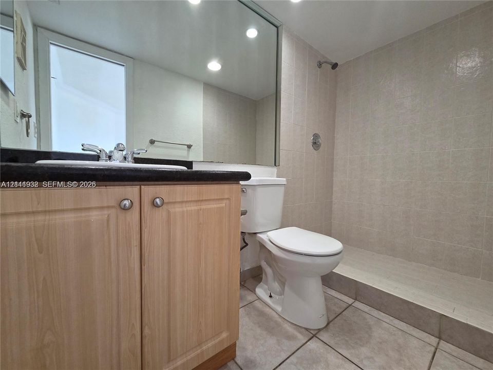 Bathroom featuring a wood-finish vanity with a dark stone-top counter, large wall-mounted mirror, ceramic tile flooring, and a walk-in shower with neutral wall tiling
