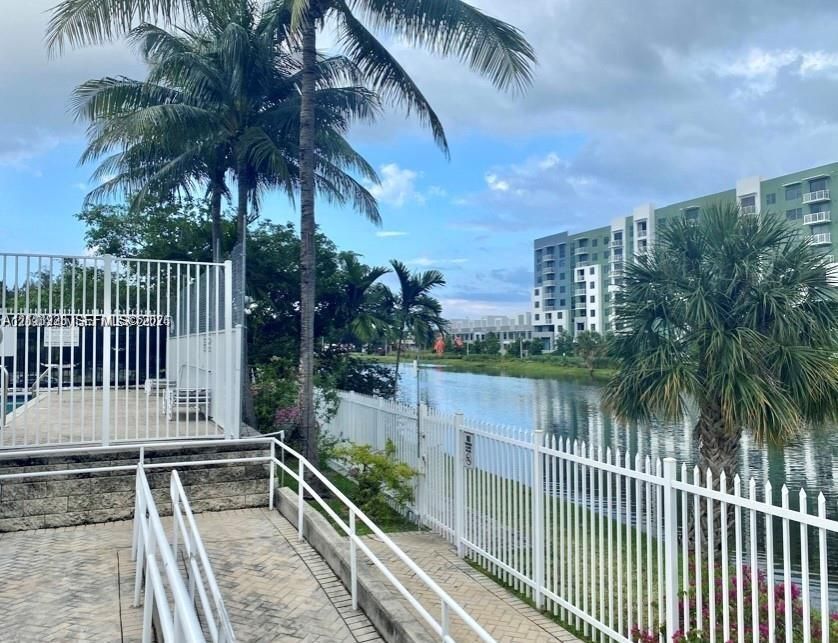 Waterfront walkway featuring white metal perimeter fencing, paved stone surfaces, and established palm trees
